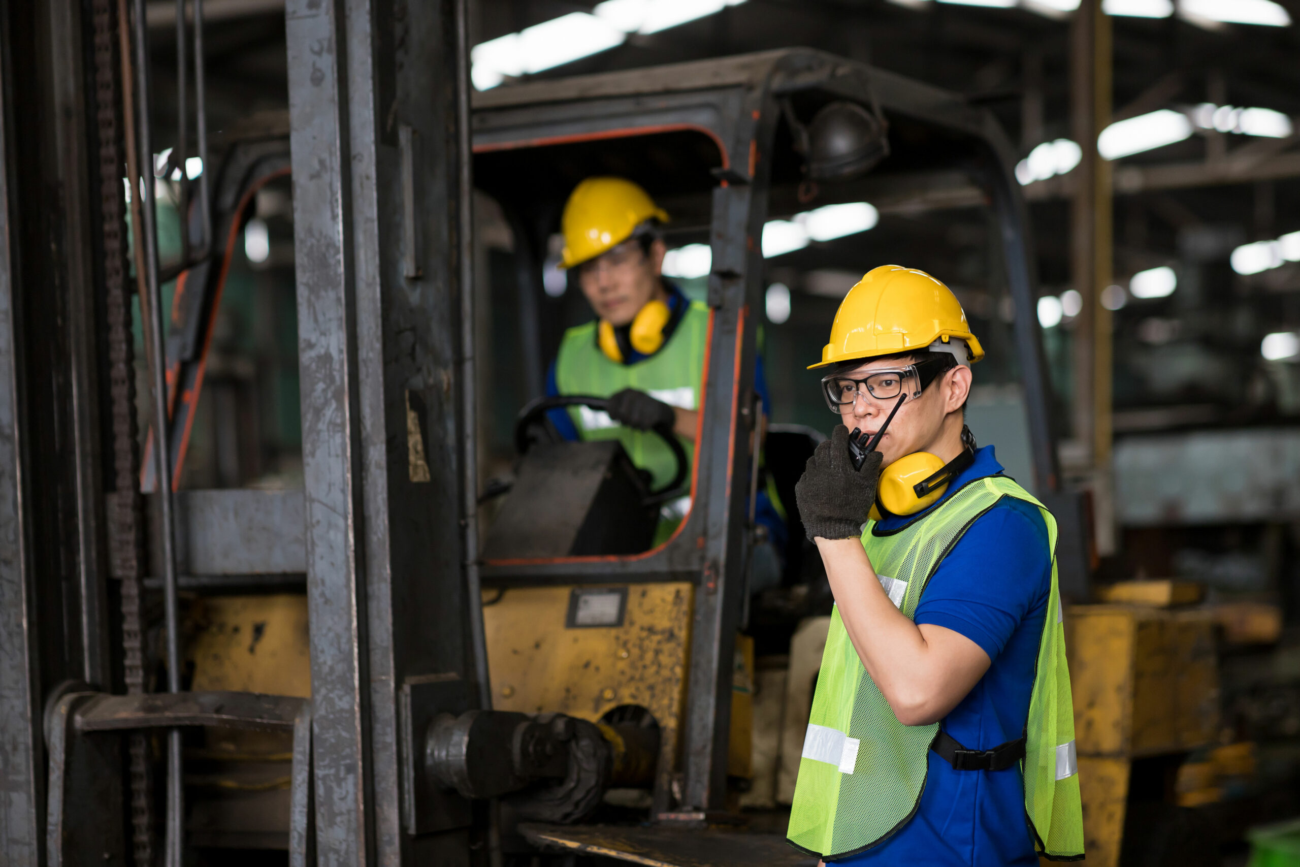 A person driving a forklift in the background as another worker stands in the foreground talking on a walkie talkie.